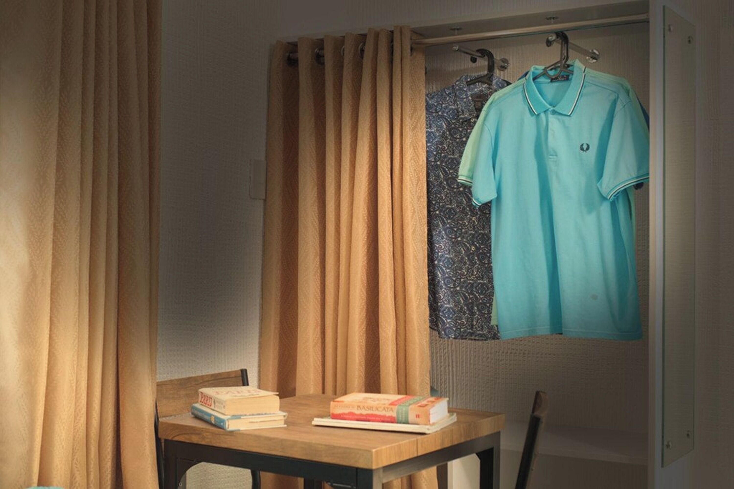 A room with a wooden table, two books, a hanging light blue shirt, and a patterned curtain.