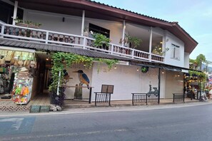 Un immeuble de deux étages doté d’un balcon, de plantes en pot et d’un commerce au rez-de-chaussée dont les volets sont fermés.