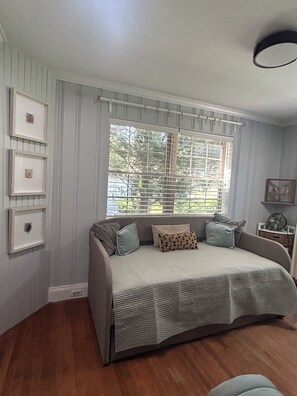 A bedroom with a bed, a window with blinds, a wall-mounted shelf, and framed pictures.