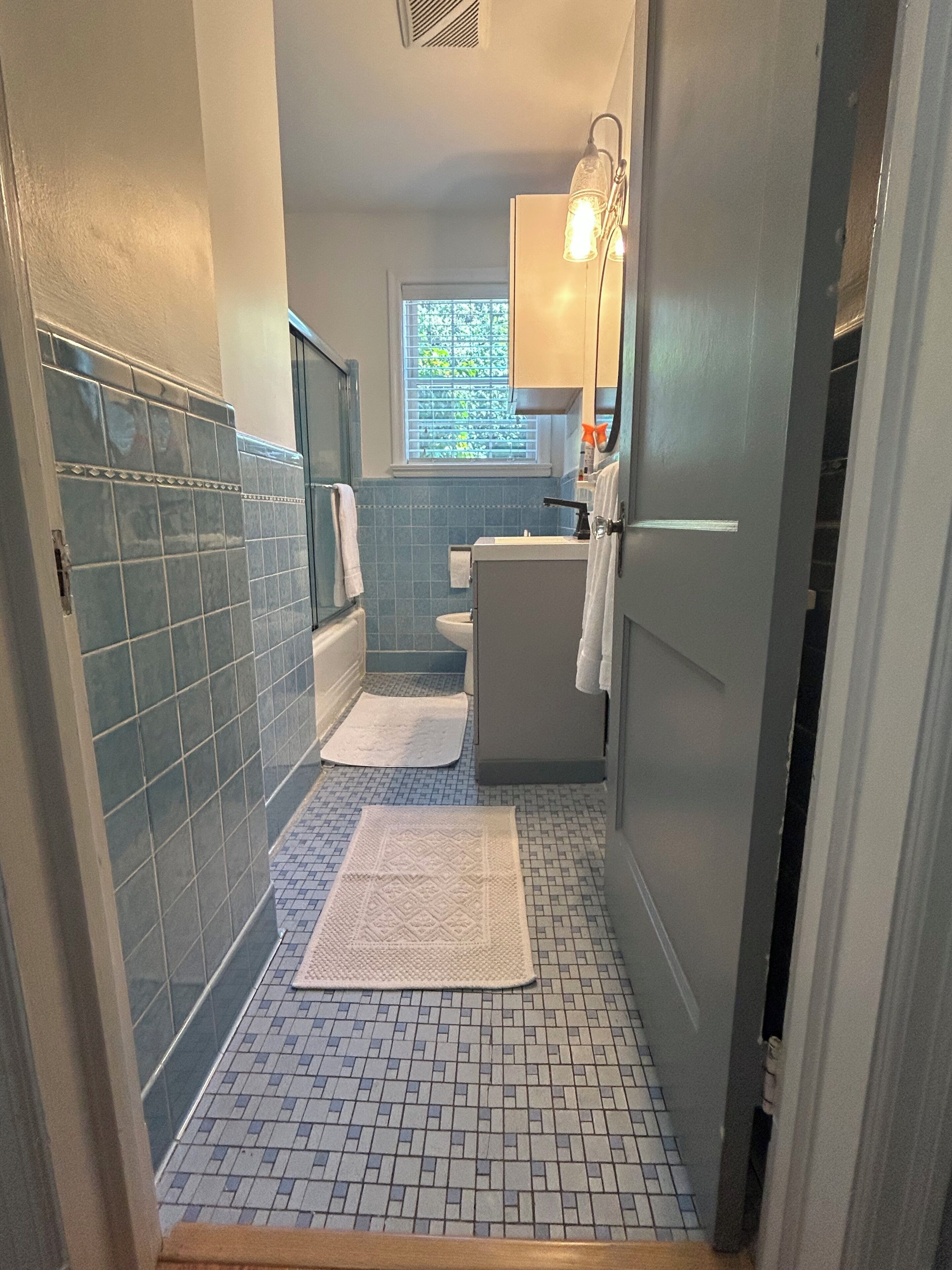 Un baño estrecho con paredes de azulejos azules, un lavamanos blanco y una ventana con persianas.
