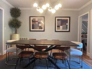 Een eetruimte met een lange tafel, zes stoelen, twee ingelijste abstracte schilderijen, een potplant en een plafondlamp.