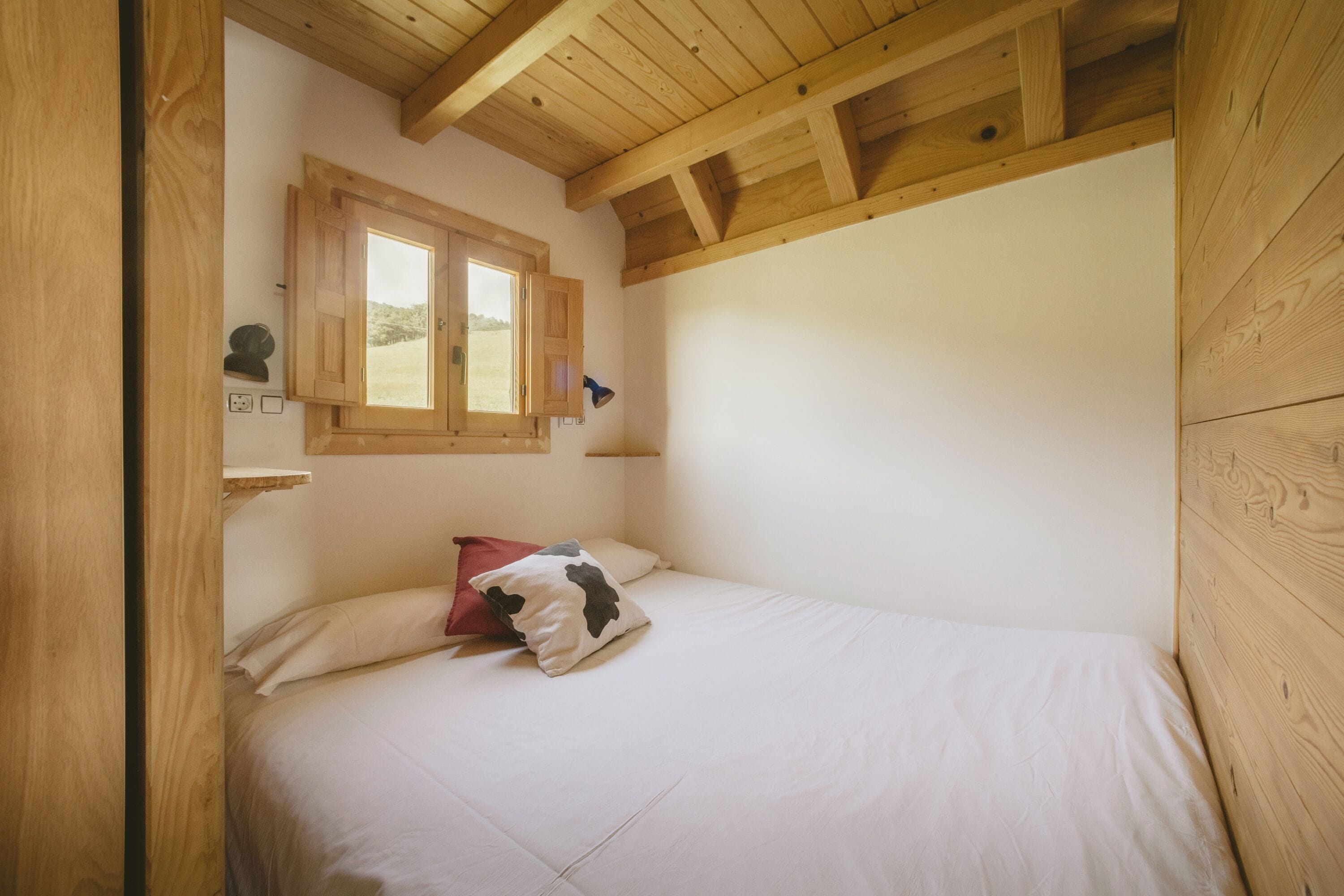 Een slaapkamer met een houten plafond, een raam met uitzicht, een bed met wit beddengoed en een rood kussen.