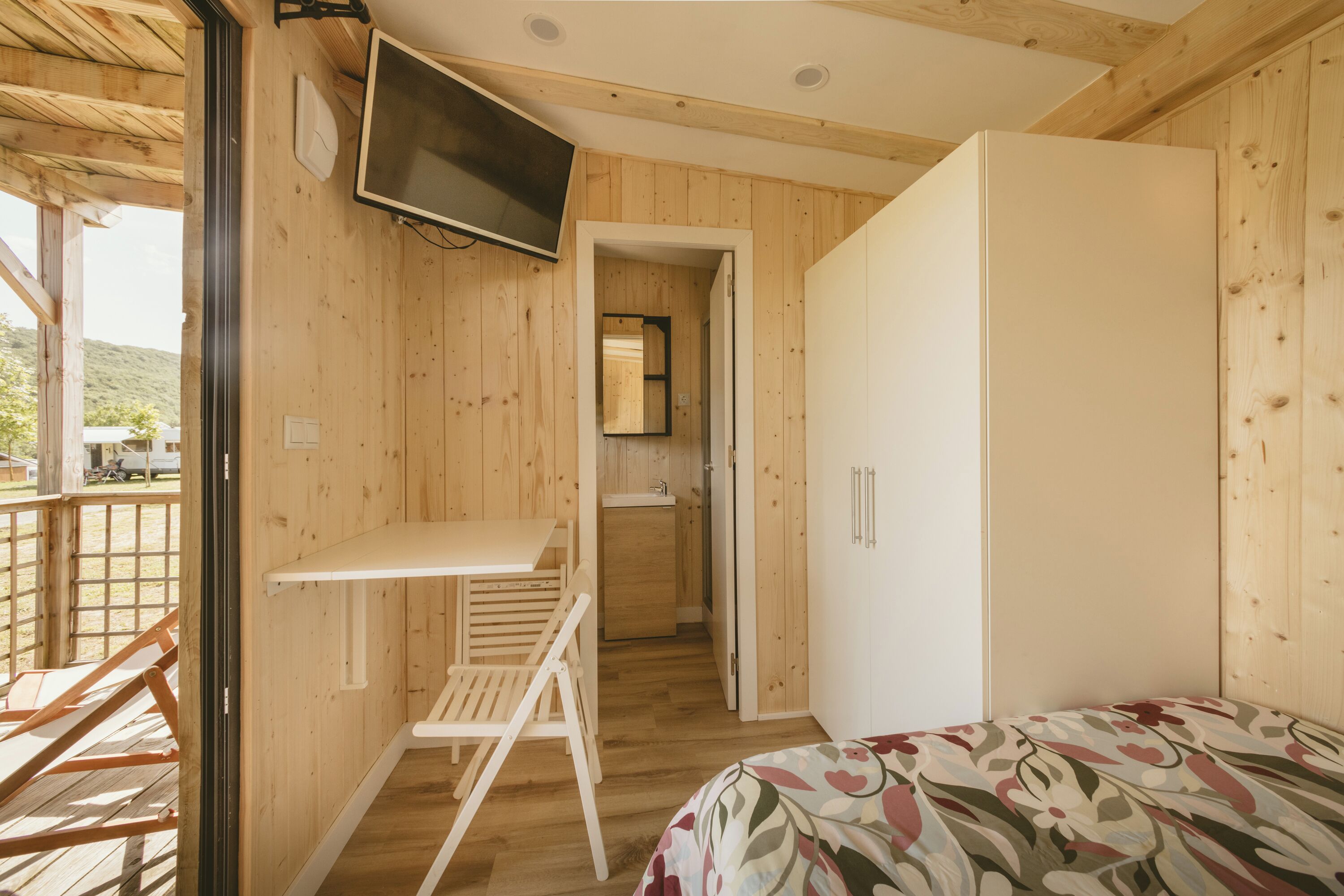 A wooden cabin with a bed, a TV mounted on the wall, a small kitchenette, and a wooden deck outside.