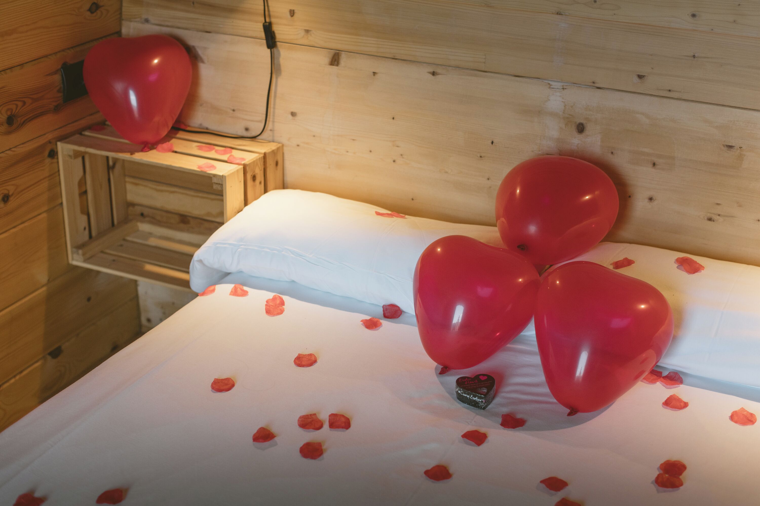 A wooden room with a bed covered in white sheets and red rose petals, and two red heart-shaped balloons.