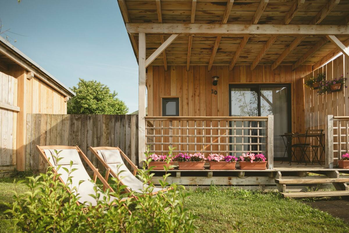 A wooden cabin with a porch, two lounge chairs, and potted flowers.