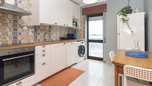 A modern kitchen with white cabinets, a built-in oven, a washing machine, and a dining area with a wooden table and chairs.