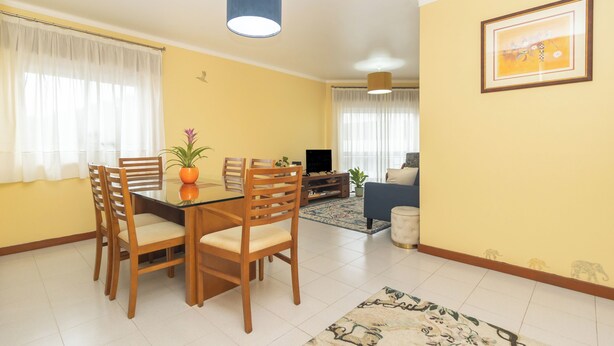 A dining area with a glass table and wooden chairs, a living room with a sofa and TV, and a yellow wall with a framed picture.