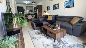 Een moderne woonkamer met een grijze bank, een salontafel en een televisie. Er staan planten, een vloerkleed en kunstwerken aan de muren.