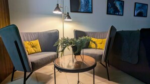 Een moderne woonkamer met twee grijze banken, gele kussens, een ronde salontafel en een vloerlamp. Er hangen drie ingekaderde kunstwerken aan de muur.