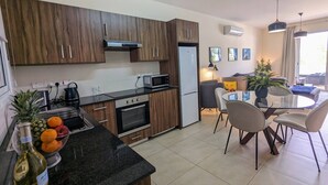 Een moderne keuken met houten kastjes, een ingebouwde oven en een magnetron. Er is een eethoek met een glazen tafel en stoelen.