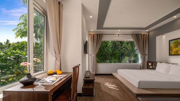 A modern hotel room with a large bed, a desk with a chair, a TV, and a view of lush greenery.
