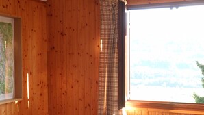 A wooden room with a bed, a window with curtains, a lamp, and a painting on the wall.