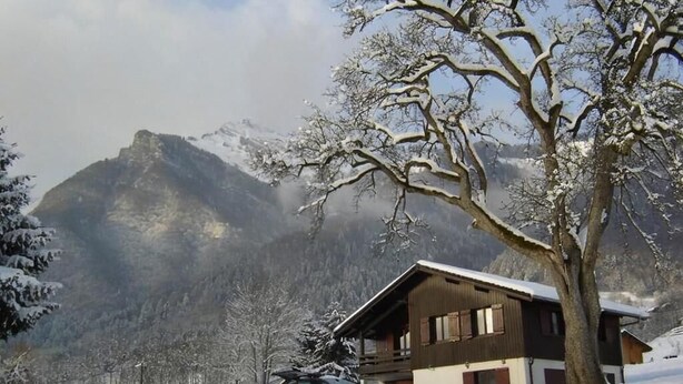 A wooden cabin with a sloped roof, surrounded by snow-covered trees and mountains in the background.