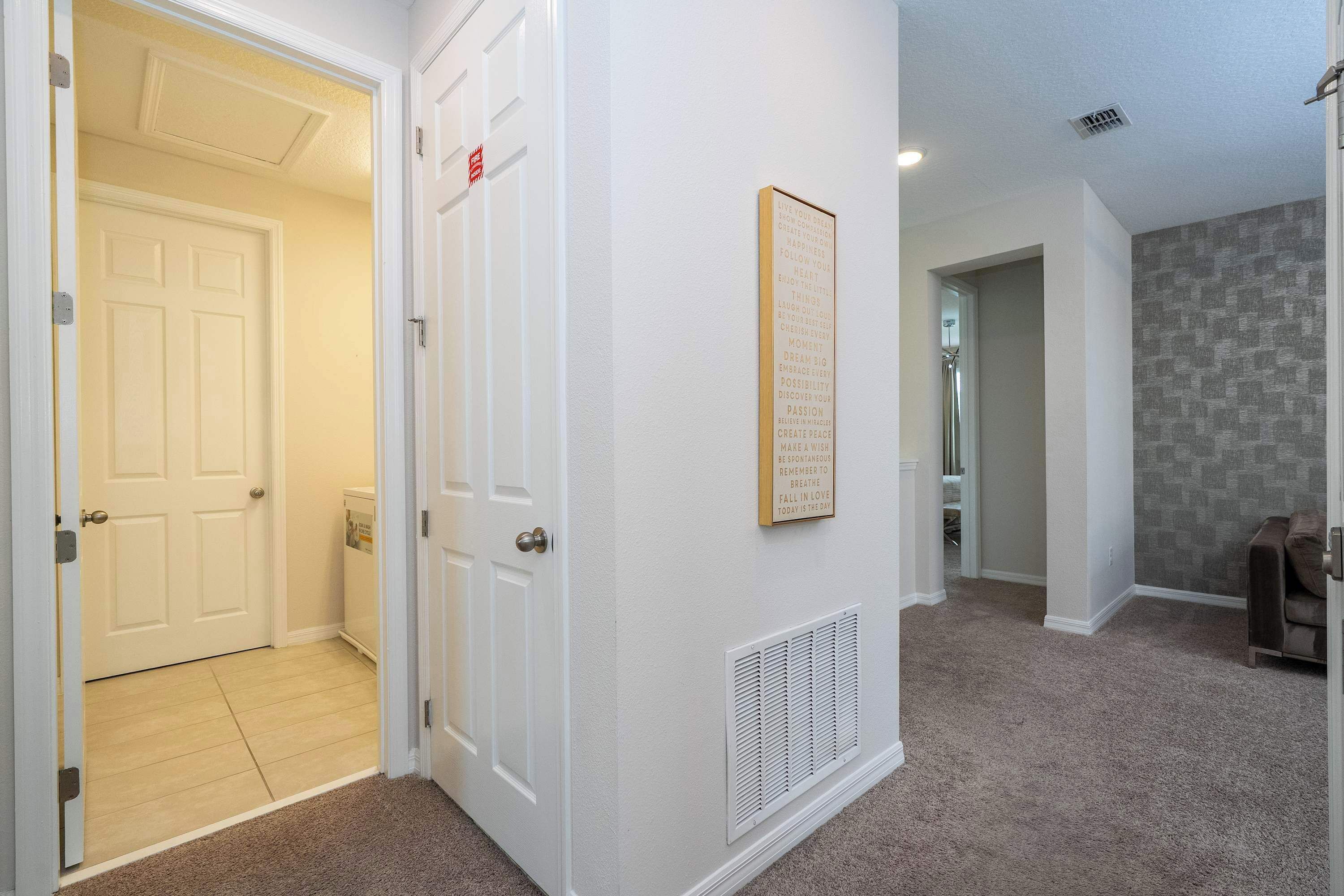 A hallway with white walls, a framed inspirational quote, a vent, and a door leading to another room.