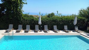 A rectangular swimming pool with clear blue water, surrounded by lounge chairs and umbrellas, with a view of the sea in the background.