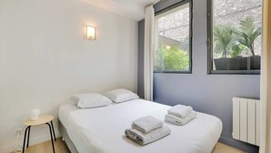 Une chambre moderne avec un grand lit, du linge de lit blanc, une petite table de chevet et deux fenêtres donnant sur un paysage verdoyant.