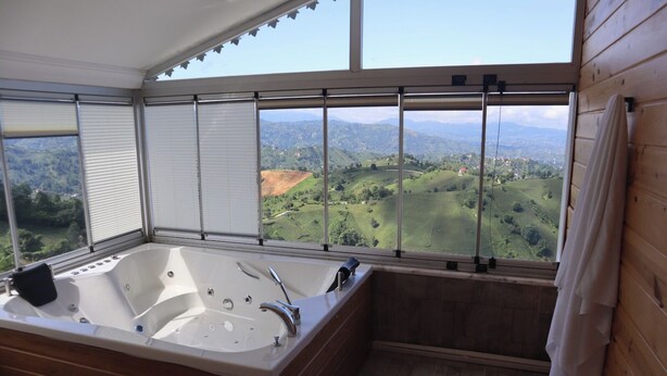 Un baño con una bañera grande, suelo de madera y vistas a un paisaje verde a través de amplios ventanales.