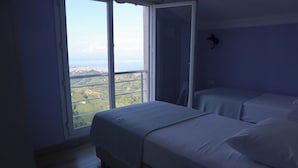 Un dormitorio con dos camas, un ventanal con vistas a un paisaje urbano y una lámpara fijada a la pared.