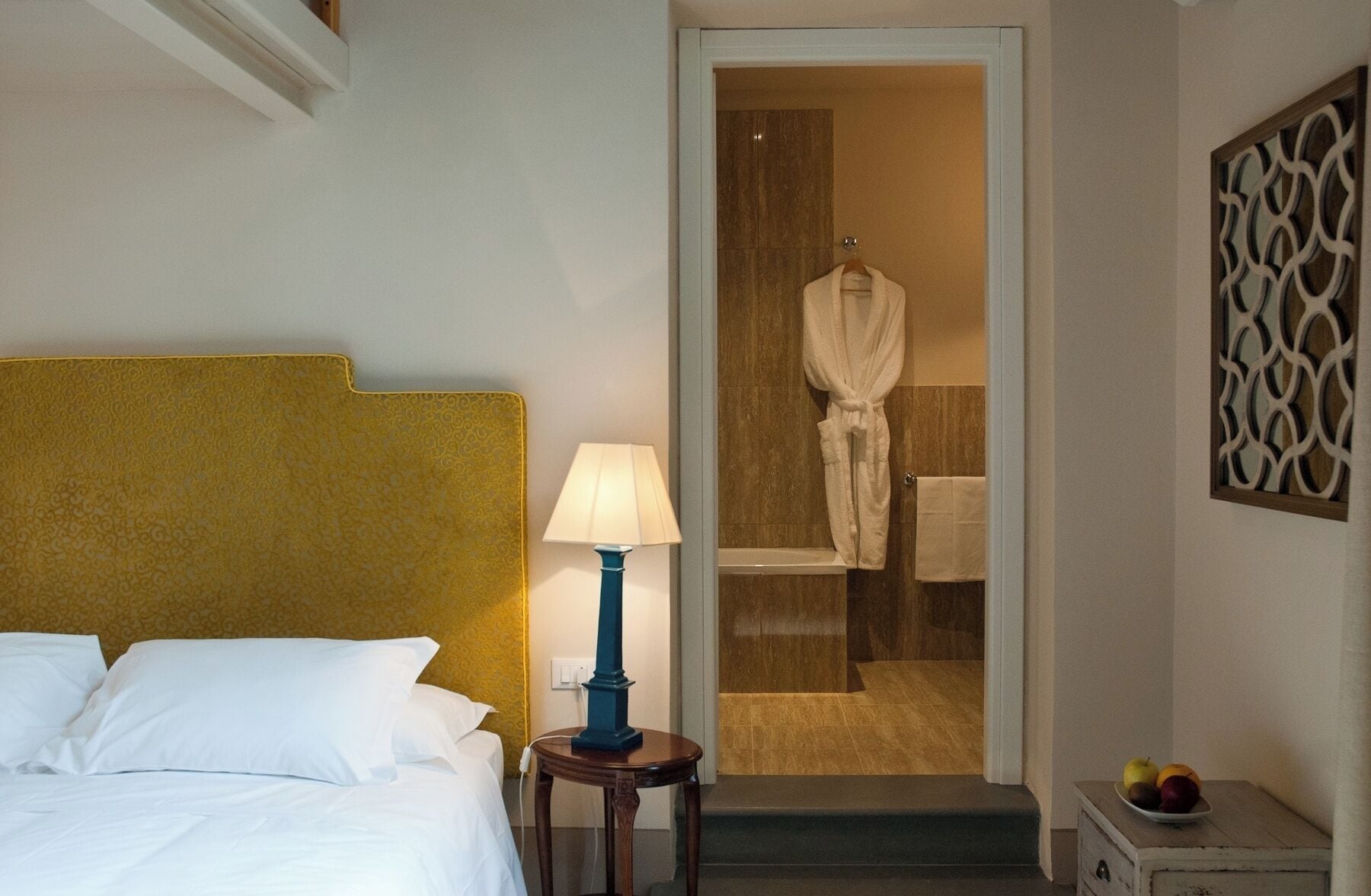 Una camera d'albergo con un letto, una lampada da comodino, un accappatoio appeso alla porta e una ciotola di frutta sul tavolino.