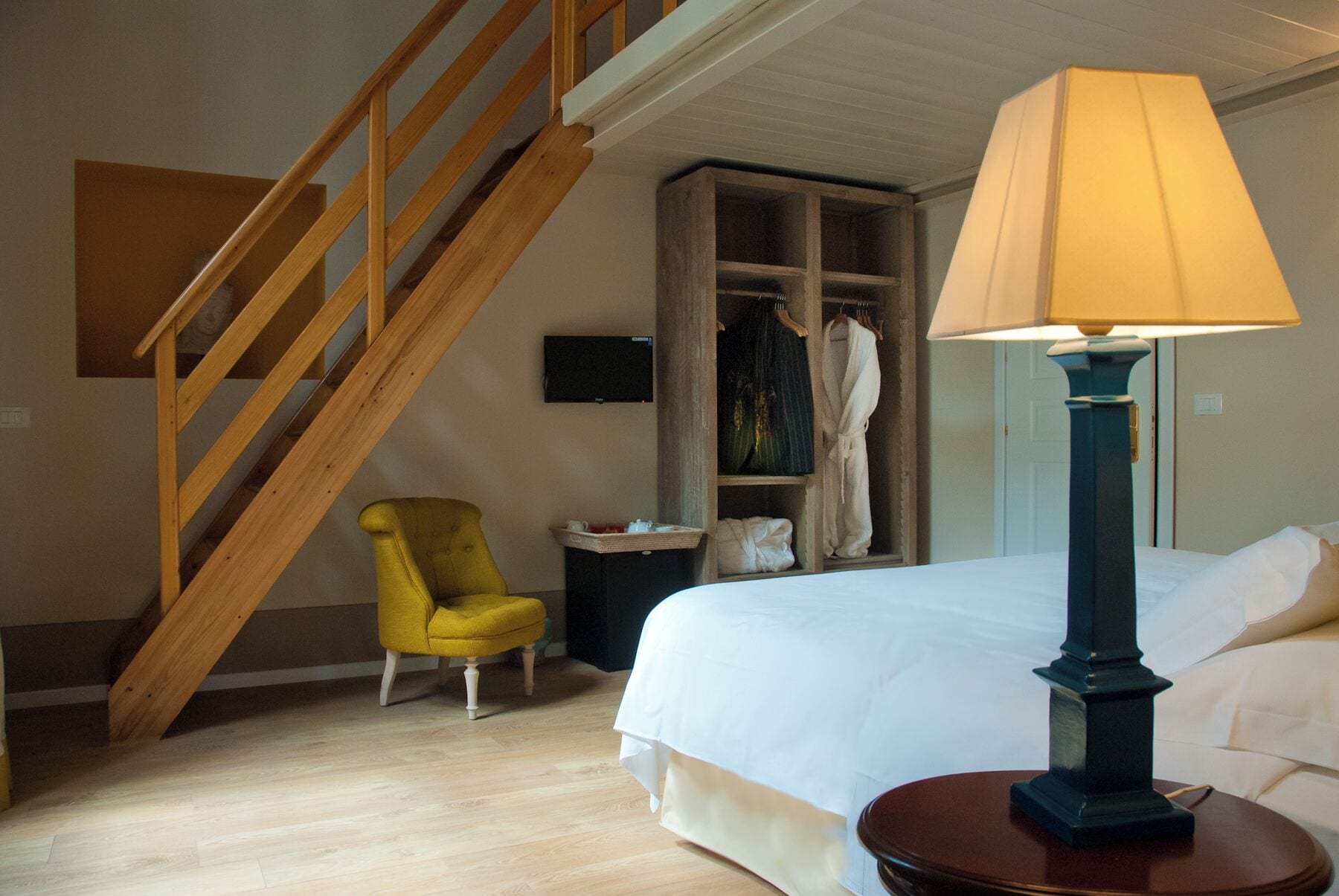 A bedroom with a wooden staircase, a yellow armchair, a bedside table with a lamp, and a wardrobe with clothes and a robe.