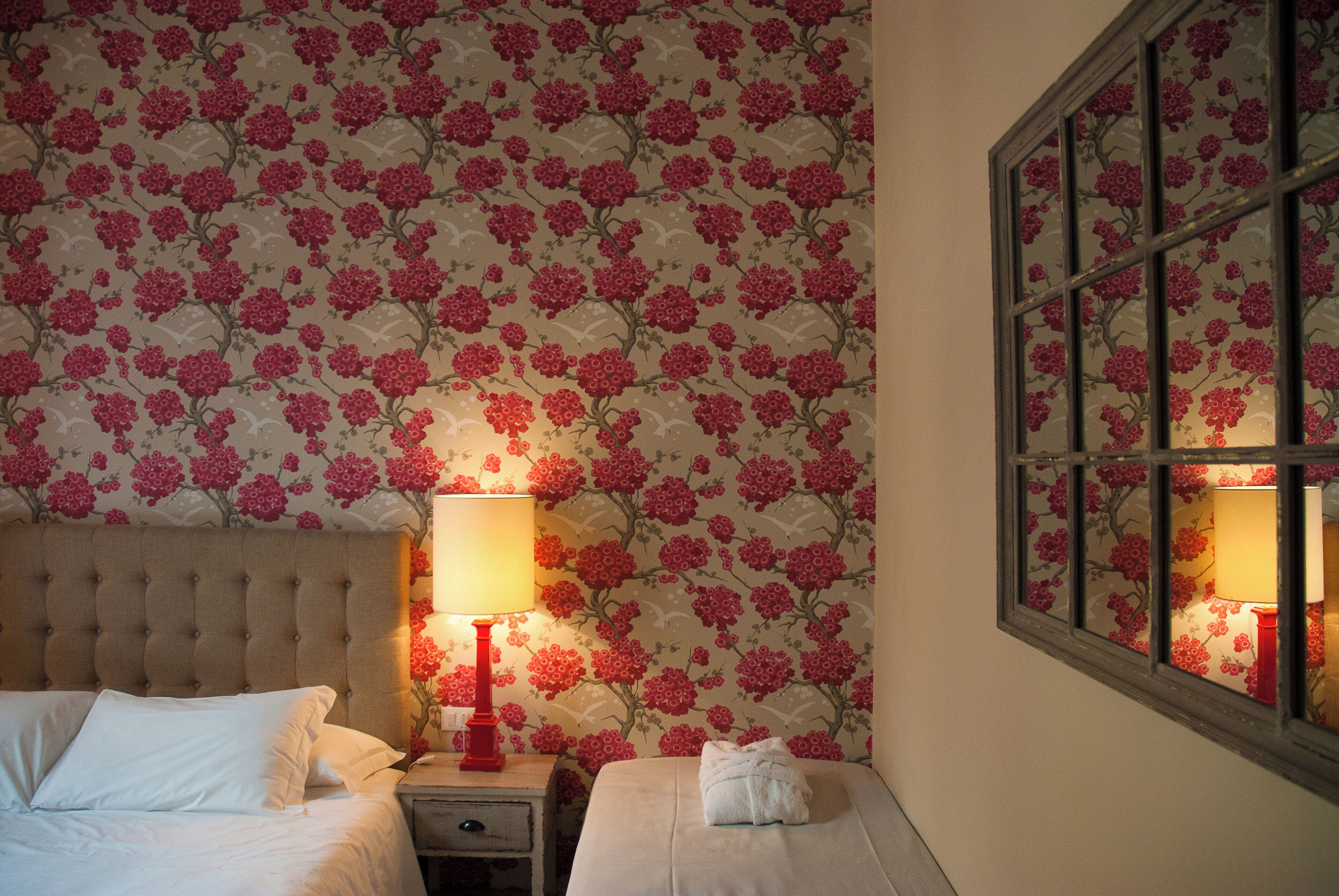A hotel room with a floral wallpaper, a tufted headboard, two beds, a nightstand with a red lamp, and a window with a view of the outside.