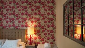 A hotel room with a floral wallpaper, a tufted headboard, two beds, a nightstand with a red lamp, and a window with a view of the outside.