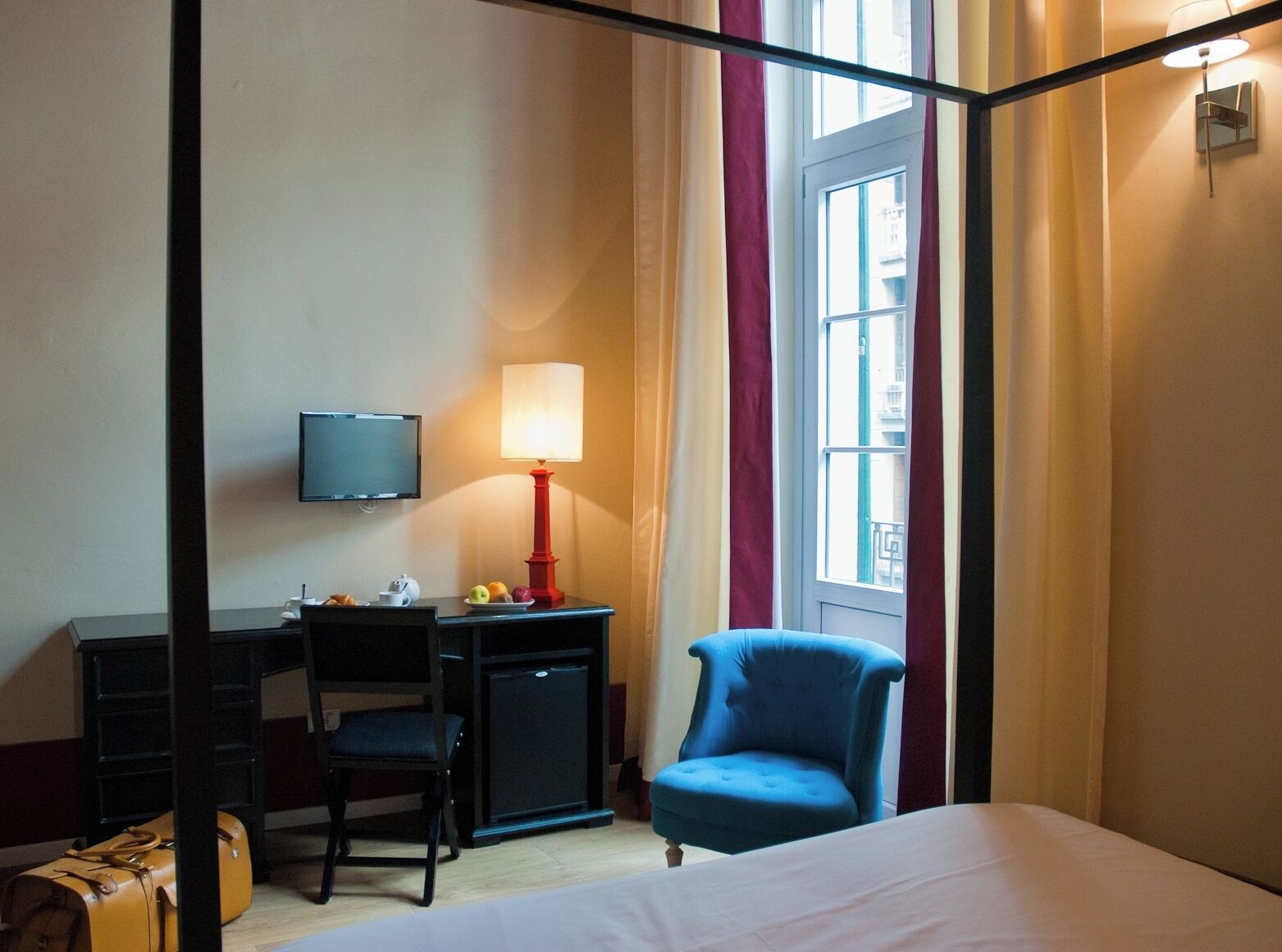 A hotel room with a bed, a desk with a chair, a television, a lamp, and a window with curtains.