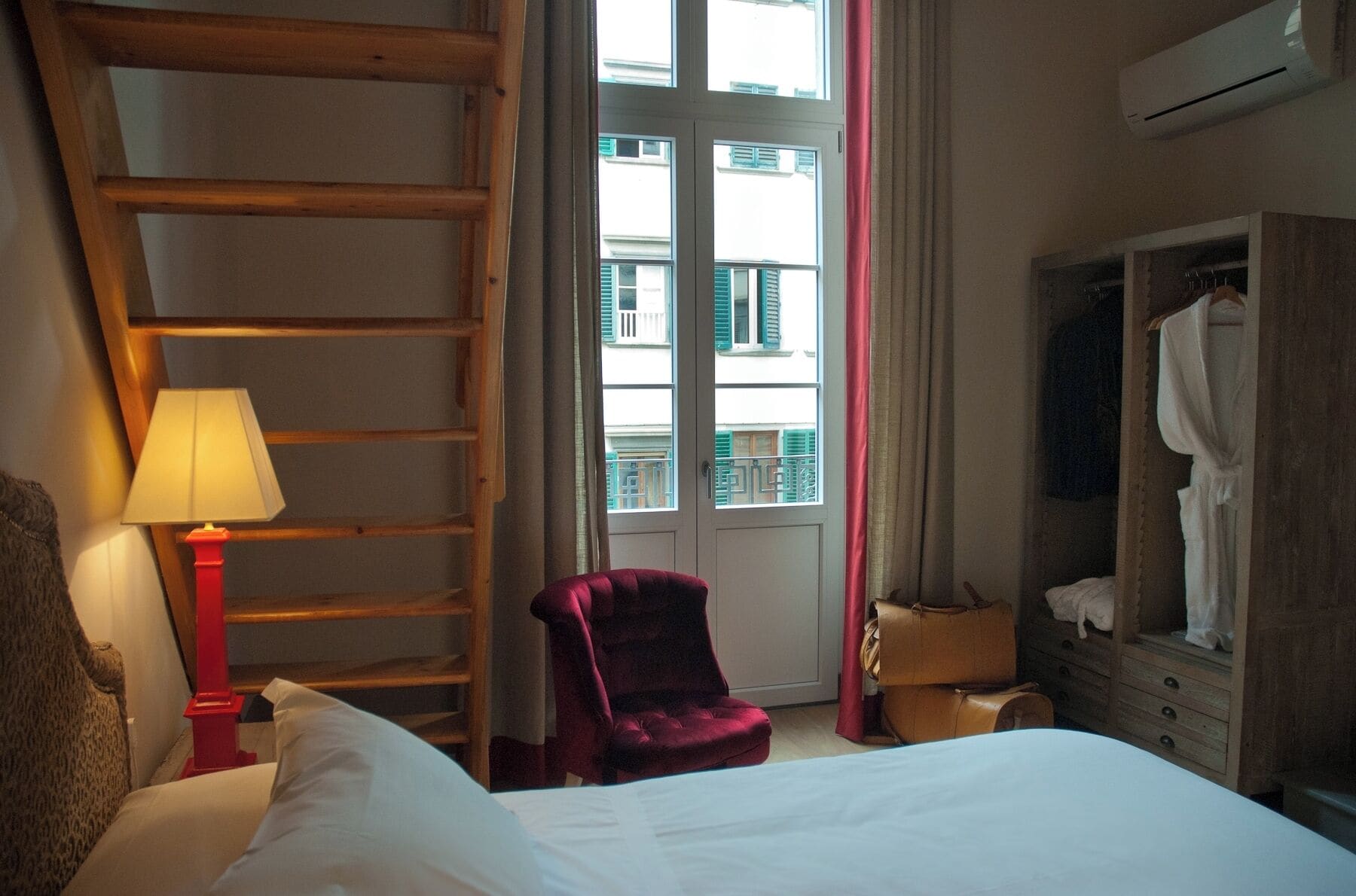 A hotel room with a bed, a red lamp, a wooden ladder, a chair, a wardrobe, and a window with a view of buildings.