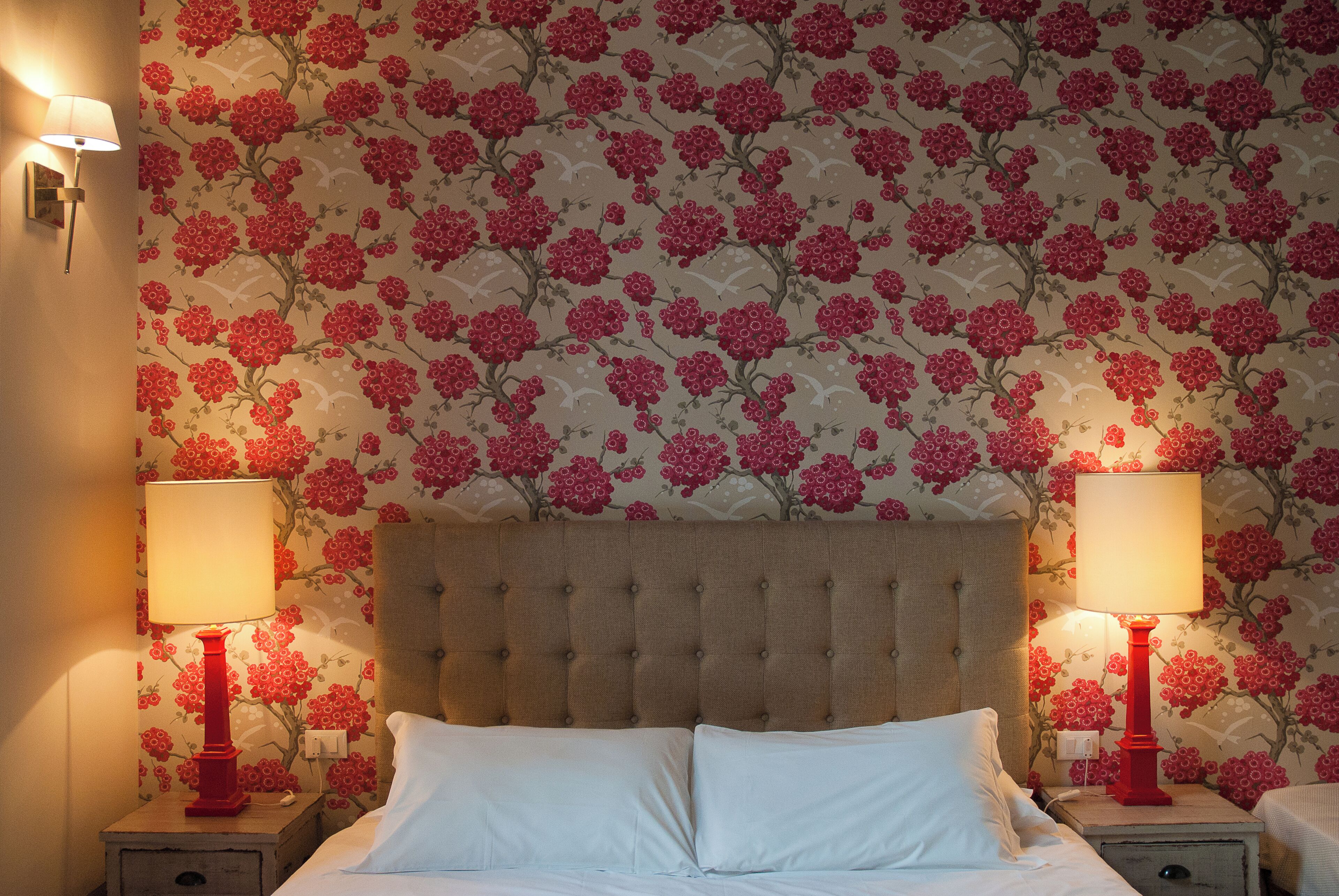 Quarto com cabeceira estofada, duas mesas de cabeceira com abajures e papel de parede floral.