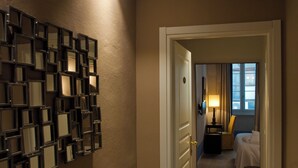 Un pasillo de hotel con una pared de cuadros enmarcados, una puerta que da a una habitación con cama y mesita de noche, y una ventana con cortinas.