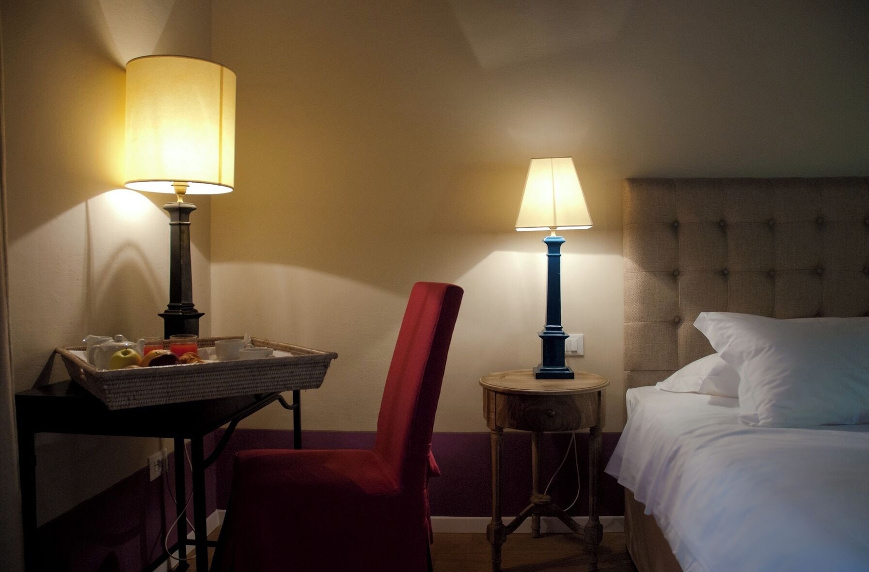 A hotel room with a bed, two bedside lamps, a red chair, and a small table with a basket containing a teapot, cups, and a fruit bowl.