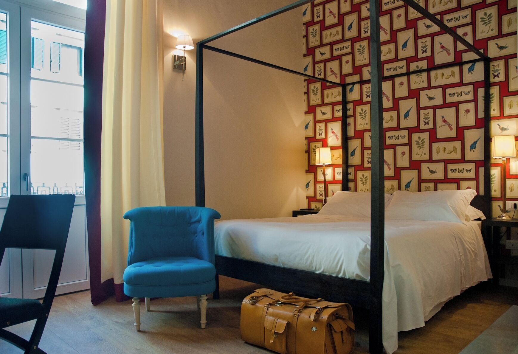 Ein Zimmer mit einem Himmelbett, einem blauen Sessel, einem braunen Koffer und einer Wand mit gerahmten Vogelbildern.