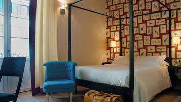 Ein Zimmer mit einem Himmelbett, einem blauen Sessel, einem braunen Koffer und einer Wand mit gerahmten Vogelbildern.