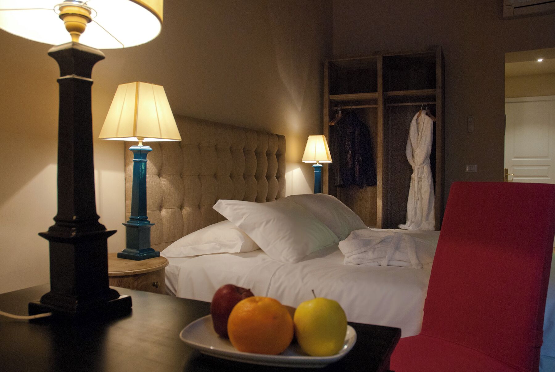 Una habitación de hotel con una cama, dos lámparas de noche, una bata colgada en la puerta y un plato de frutas sobre una mesa.