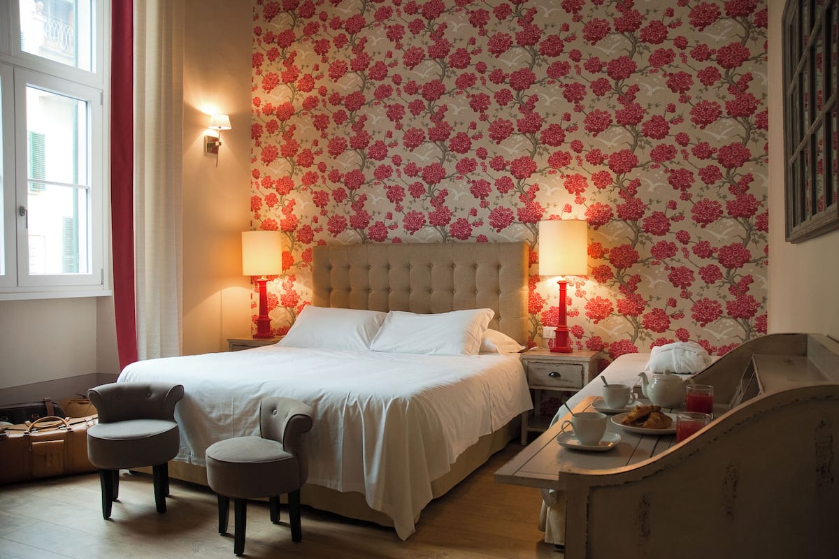 Un dormitorio ordenado con una cama grande, dos mesitas de noche con lámparas, una pequeña mesa con tetera y tazas, y una silla.