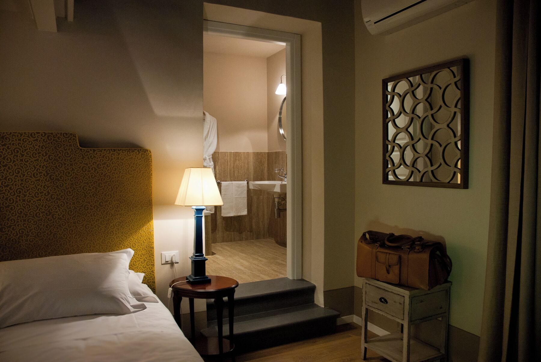 A hotel room with a bed, a nightstand with a lamp, a suitcase on a small table, and a bathroom visible through an open door.