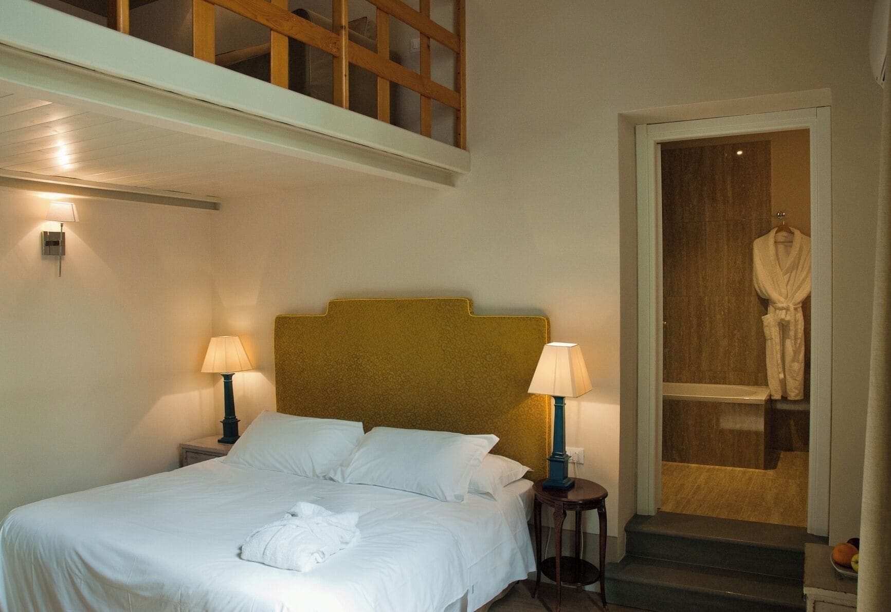A hotel room with a bed, two bedside lamps, a robe hanging on a door, and a staircase leading to an upper level.