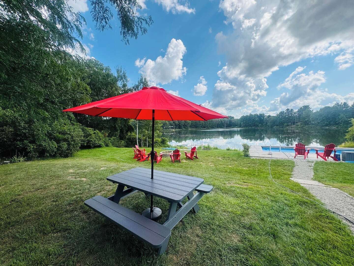 Ein Picknicktisch mit rotem Sonnenschirm, umgeben von roten Stühlen, in der Nähe eines Schwimmbades und eines Sees.
