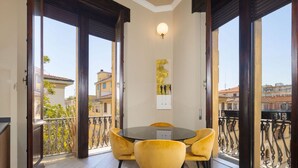 Un espace repas avec une table ronde et des chaises jaunes, de grandes fenêtres donnant sur des immeubles et un luminaire fixé au mur.