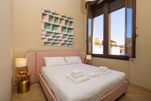 A hotel room with a bed, pillows, a wall art piece, a window, and a nightstand with a lamp. - Tuscan Refined Retreat (Firenze)
