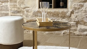 A round table with a gold frame and a tray, placed on a white rug. In the background, there is a stone wall with a shelf containing decorative items.