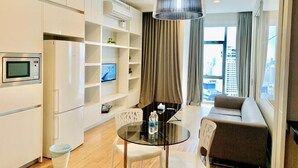 Une chambre d’hôtel moderne dotée d’une kitchenette, d’un coin repas avec une table en verre, d’un canapé et d’une grande fenêtre offrant une vue sur la ville.