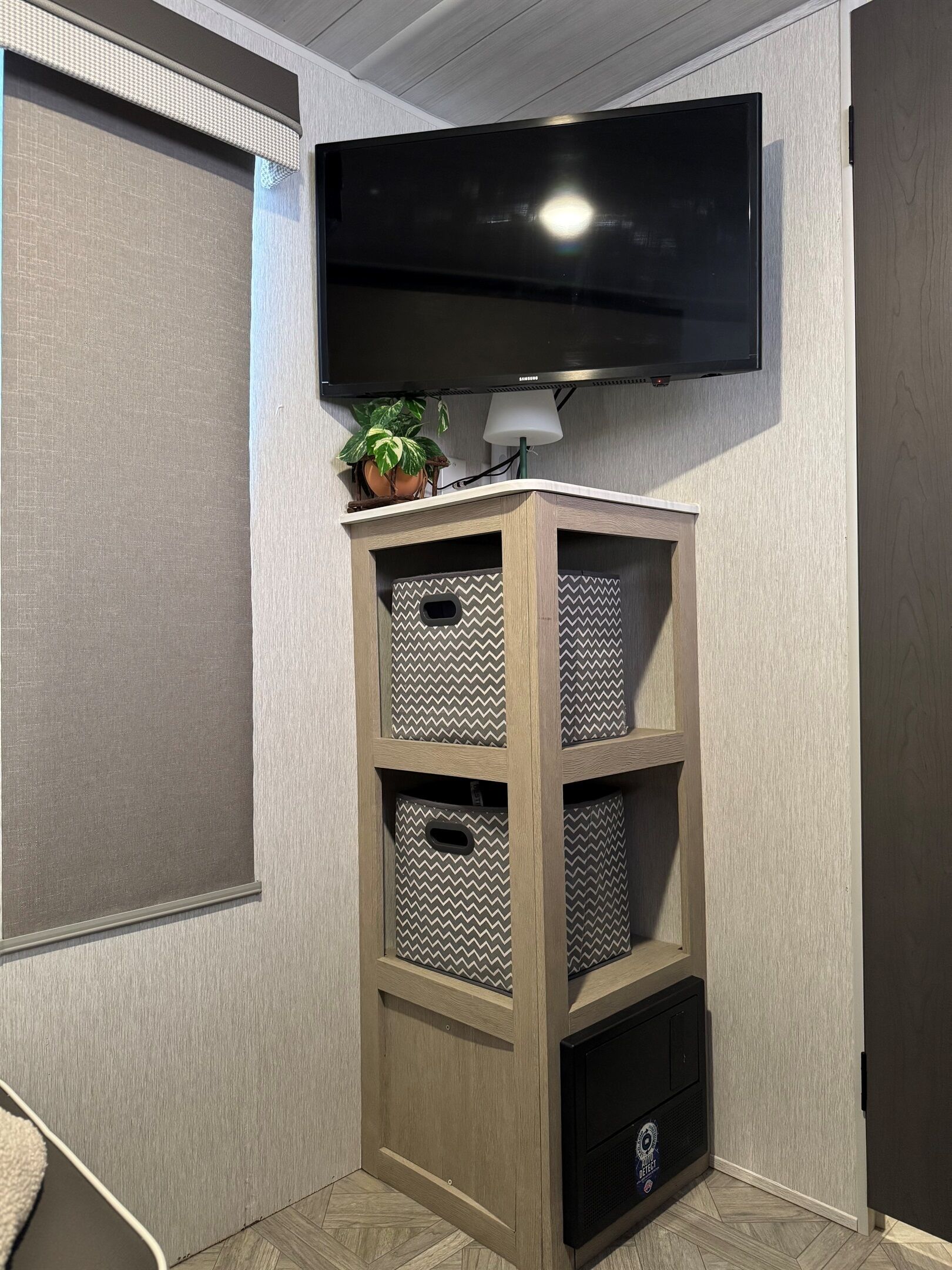 Een hoekappartement met een wandgemonteerde televisie, houten planken met decoratieve opbergmanden, een potplant en een lamp.