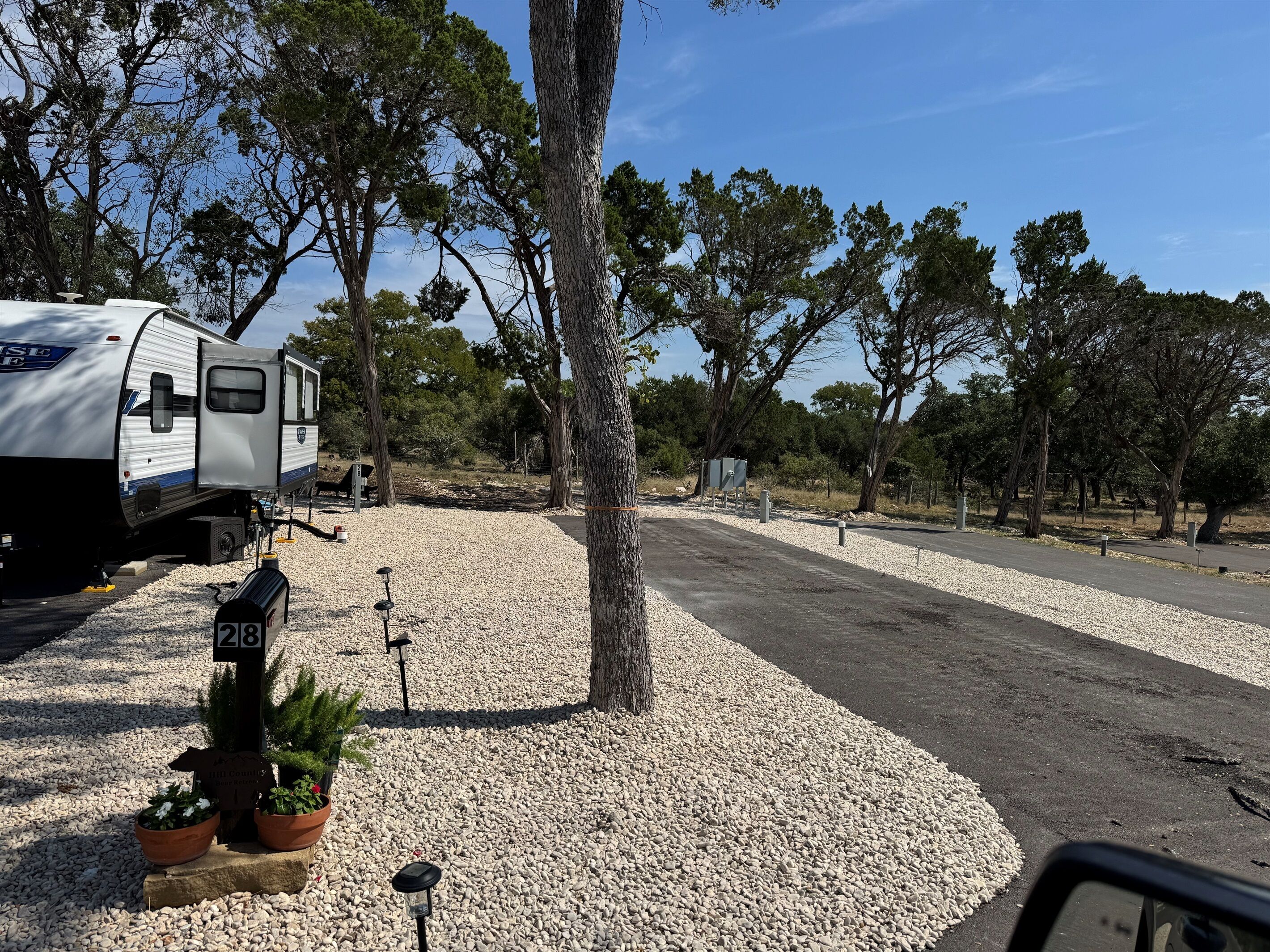 A campsite with a white RV parked on gravel, surrounded by trees and a paved path.