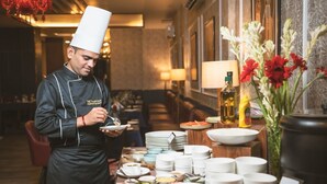 Un chef con uniforme negro y gorro blanco sirve comida en una mesa de buffet con diversos platos y condimentos.
