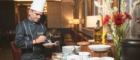 Un chef con uniforme negro y gorro blanco sirve comida en una mesa de buffet con diversos platos y condimentos.
