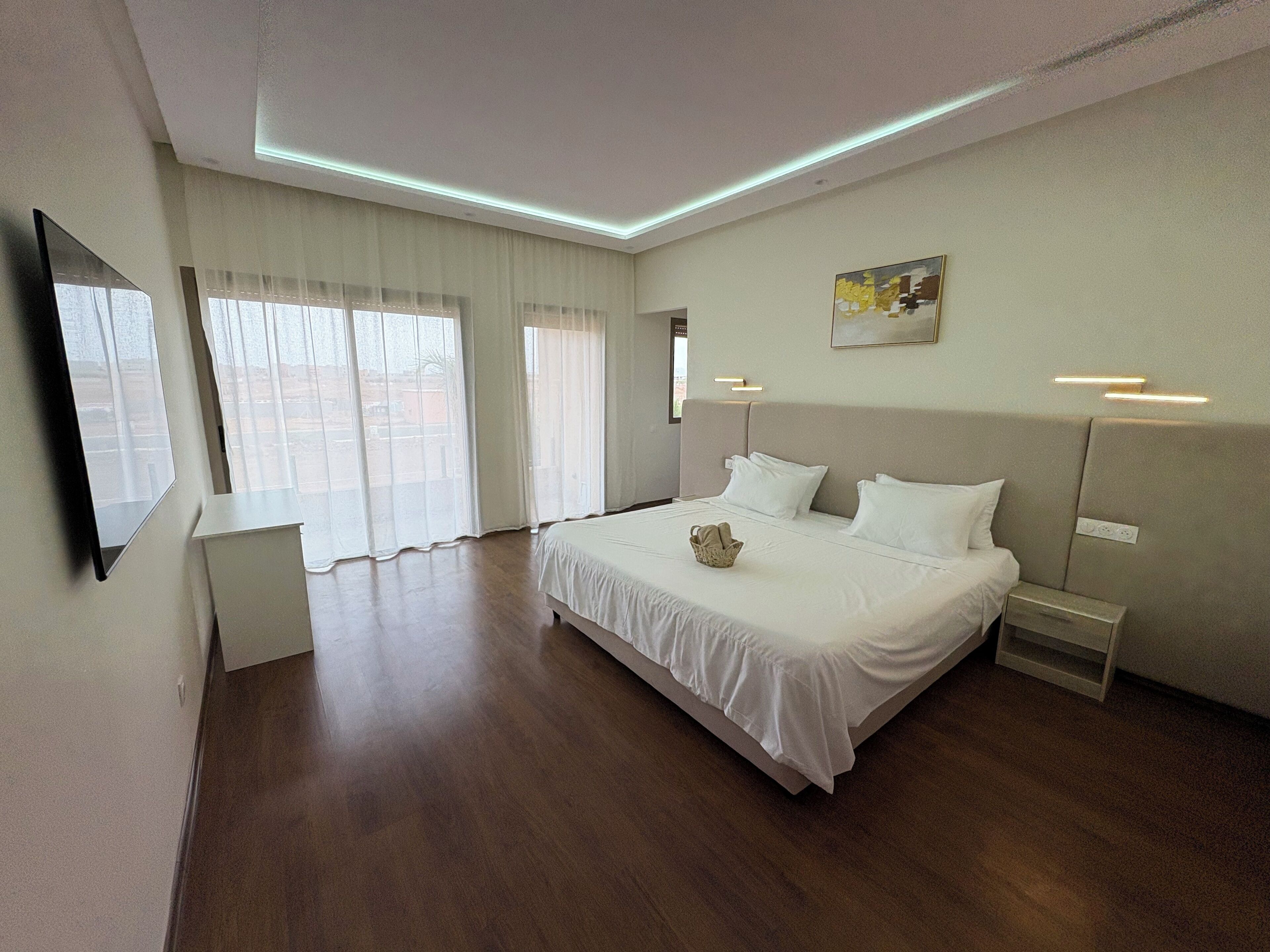Une chambre d’hôtel moderne dotée d’un grand lit, d’un téléviseur à écran plat fixé au mur et d’une grande fenêtre avec des rideaux légers.