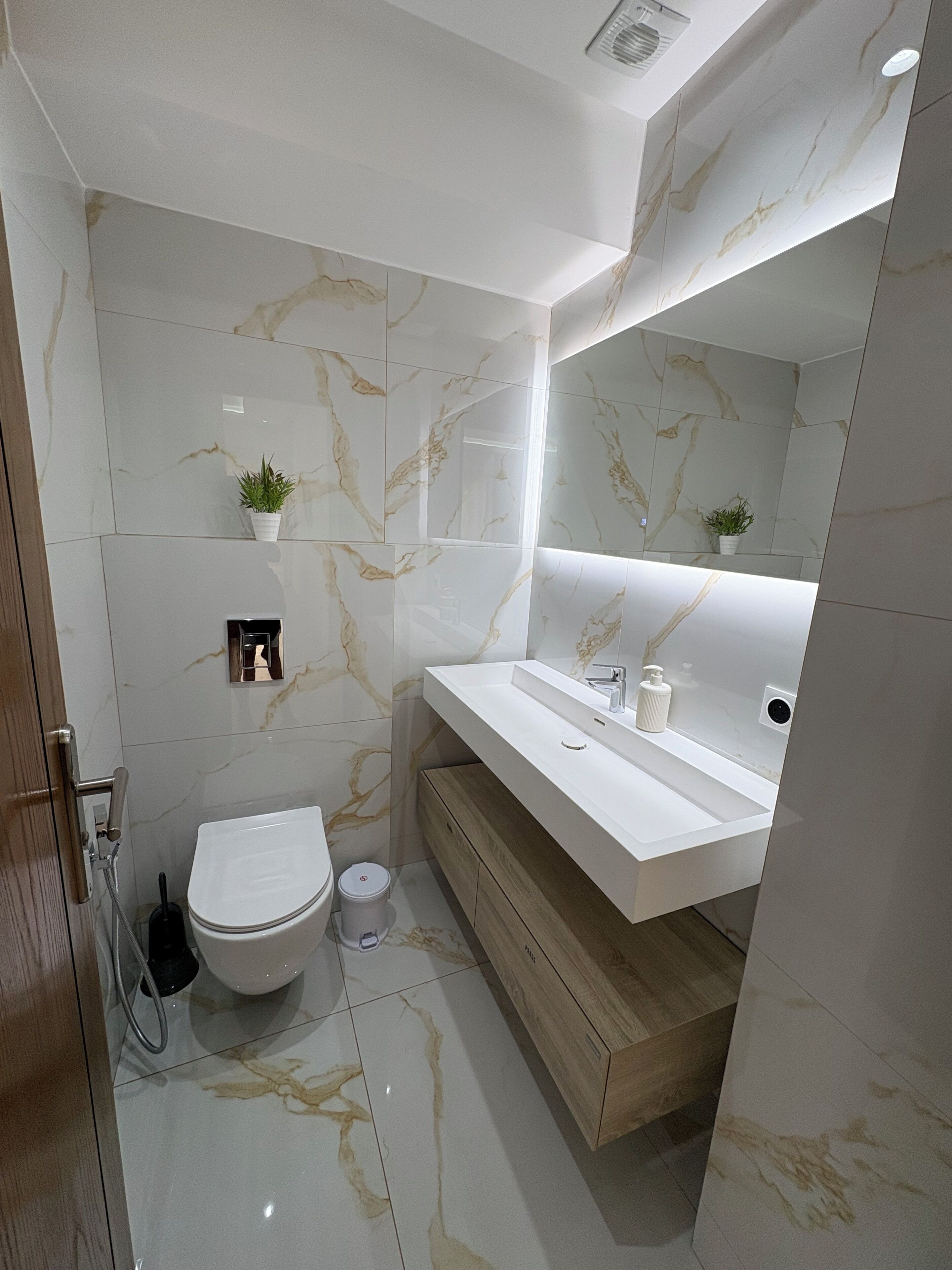 Une salle de bain moderne avec un sol en marbre, des toilettes blanches, un meuble-lavabo en bois et une petite plante en pot.