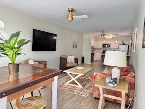A living room with a TV, a ceiling fan, a dining table, and a kitchen in the background. - Sea Oats C203 2 bedroom / 2 bathroom  pet friendly Complete remodel (Gulf Shores)