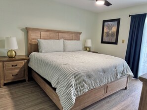 A bedroom with a wooden bed, two bedside tables with lamps, a nightstand, a framed picture on the wall, and a window with curtains. - Sea Oats C203 2 bedroom 2 bathroom Pet-Friendly Complete remodel (Gulf Shores)